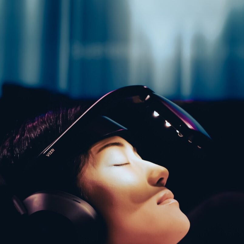 Person lying back wearing an oversized VR-style headset in a dark room, illuminated by soft blue and purple light.
